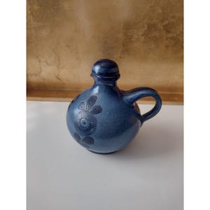 Könnte beinhalten: Ein blauer Keramikkrug mit Henkel und Deckel. Der Krug hat ein Blumendesign auf der Vorderseite. Der Krug steht auf einer weißen Oberfläche mit goldenem Hintergrund.