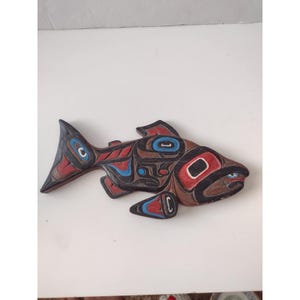 Könnte beinhalten: Eine geschnitzte Holzfischskulptur mit einem indianischen Design. Der Fisch ist hauptsächlich braun mit roten, blauen und schwarzen Akzenten. Das Kunstwerk weist geometrische Formen und Muster auf und ist wahrscheinlich ein Wandbehang oder ein dekoratives Stück.