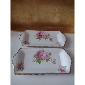 May include: Two rectangular white porcelain serving trays with scalloped edges and gold-toned trim. Each tray features a central cluster of pink roses with green leaves and smaller rose accents in the corners of the white surface.