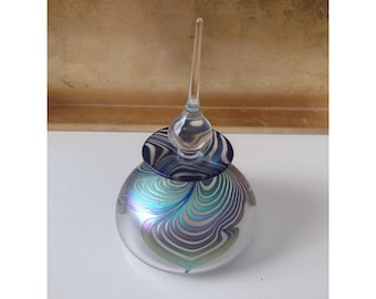 May include: A round glass perfume bottle featuring an iridescent finish with swirling teal, purple, and silver patterns. The bottle is topped with a clear, pointed glass stopper that has a dark blue and purple circular base with swirling lines.