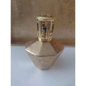 May include: A faceted, metallic gold fragrance lamp with a textured, diamond-like body. The top features a cylindrical, perforated gold-tone metal cap with a visible wick assembly inside, designed for air purification and home fragrance diffusion.