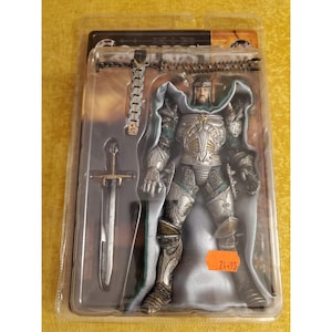 Op de afbeelding: Een in plastic verpakte actiefiguur van een ridder in zilverkleurig harnas met een groene cape, inclusief een zwaard en een kruisvormig accessoire. De verpakking bevat een feloranje sticker met de handgeschreven tekst 24.95.