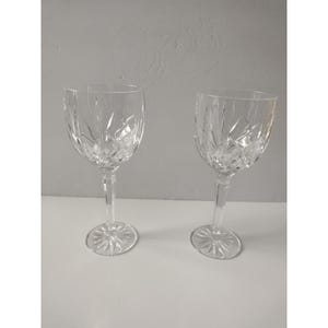 Op de afbeelding: Twee wijnglazen van helder kristal met een verticaal geslepen patroon op de kelken en een stervormig ontwerp op de ronde voeten. Elk glas heeft een slanke steel en een afgeronde kelk, wat zorgt voor een reflecterend, gefacetteerd oppervlak.