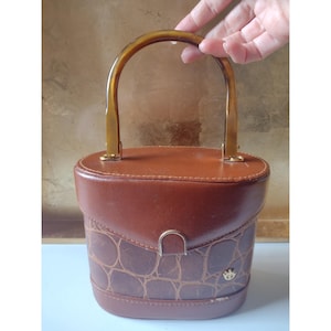 May include: A structured brown handbag with a smooth top section and a textured, crocodile-embossed lower panel. It features a rounded amber-colored plastic handle, a gold-toned metal clasp closure, and a small gold-toned emblem on the front.