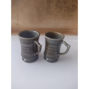 May include: Two gray ceramic mugs with handles. The mugs have a cylindrical shape with a decorative band of small, raised dots and lines around the middle. The handles are curved and attached to the side of the mugs. The mugs are on a white surface.