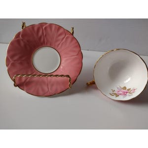 Könnte beinhalten: Rosa Teetasse und Untertasse mit Goldrand. Die Untertasse hat einen gewellten Rand und wird auf einem goldfarbenen Ständer präsentiert. Die weiße Tasse hat ein rosa Rosenmuster und Goldverzierungen.