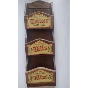 May include: A vertical wall-mounted wooden organiser featuring three tiered pockets. Each pocket has a cream-coloured label with red text reading Letters, Bills, and Misc, accented by small floral motifs. The wood has a dark brown finish.