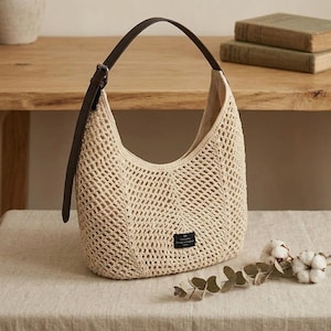 May include: A beige hobo-style handbag featuring an open-weave crochet construction. The bag includes a dark brown adjustable leather strap with a metal buckle and a small rectangular black label with white text attached to the front exterior.