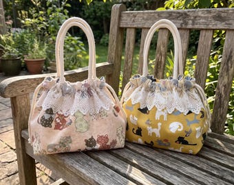 May include: Two fabric drawstring bags with white rope handles and white lace trim. The left bag is light pink with a multi-colored cat pattern. The right bag is mustard yellow with a white, black, and gray cat pattern. Both bags feature a gathered top.