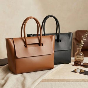 May include: Two structured tote bags, one in tan brown and one in black, featuring a smooth finish, a wide front flap, and dual top handles. The bags are positioned side by side, showcasing a clean design with a rectangular base and vertical sides.
