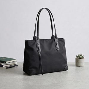 May include: A black tote bag with two long shoulder straps, featuring reinforced leather-like accents at the base corners and strap attachments. The bag has a smooth, solid black exterior with a single dangling strap detail on the front panel.
