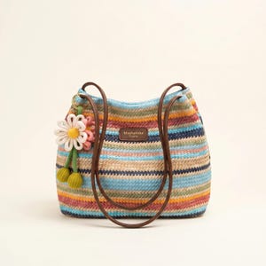 May include: A woven straw tote bag featuring horizontal stripes in shades of blue, pink, yellow, and tan. The bag has two brown leather-like shoulder straps and a brown rectangular label reading MashaKeke Original. A white flower charm hangs from a ring.