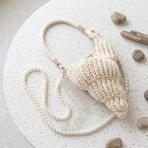 May include: A cream-colored crochet bag shaped like a croissant, featuring a braided rope handle and a long, detachable strap made of white faux pearls. The bag has a textured, knotted surface and is attached with gold-toned metal hardware.