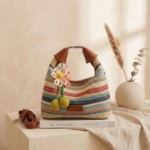 May include: A woven straw handbag featuring horizontal stripes in shades of blue, pink, yellow, and tan. The bag has brown leather-like handles, a metal clasp, and a decorative flower charm with two green pom-poms. A detachable brown strap is included.