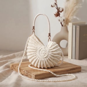 Può includere: Una borsa a mano all'uncinetto color crema a forma di conchiglia, caratterizzata da un design testurizzato a coste con un centro circolare. La borsa include un manico intrecciato fissato con hardware dorato e una lunga tracolla di perle bianche.