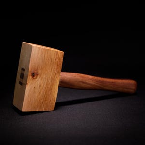 May include: A wooden mallet featuring a light-colored rectangular head with a visible wood grain and a small knot, joined to a darker, smooth, rounded wooden handle. The head includes a small, dark rectangular inlay on its side surface.