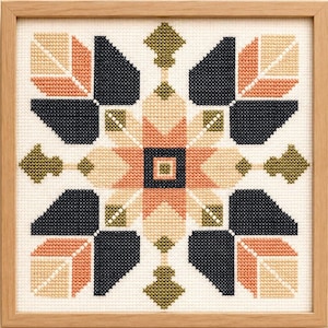May include: A square cross-stitch embroidery piece framed in light wood. The design features a symmetrical geometric pattern with a central dark blue square, surrounded by peach, cream, and olive green shapes on a white fabric background.