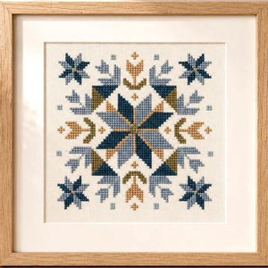 May include: A square cross-stitch embroidery featuring a symmetrical geometric star pattern in shades of navy blue, light blue, and mustard yellow, displayed in a light-toned wooden frame with a white mat board border.