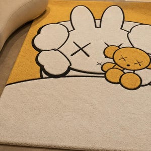 May include: A rectangular rug with a cartoon bunny and teddy bear design. The bunny is white with black outlines and cross-shaped eyes. The teddy bear is yellow with black outlines and cross-shaped eyes. The background is yellow and beige.