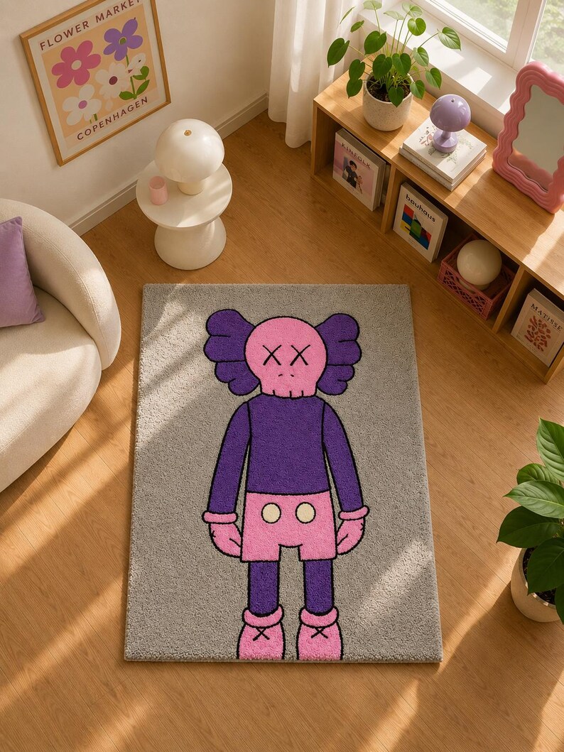 May include: A rectangular gray area rug featuring a central graphic of a cartoon character with a pink skull-shaped head, X-shaped eyes, a purple long-sleeved shirt, pink shorts with two white circular buttons, and pink shoes with cross details.