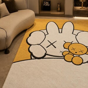 May include: A large, plush area rug featuring a cartoon character design. The rug has a white base with a yellow accent and a large, white character with black outlines and crossed-out eyes. A small, yellow teddy bear is also featured.