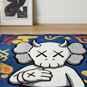 Pode incluir: Um tapete retangular com uma personagem de desenho animado branca central com chifres e olhos em forma de X sobre um fundo azul profundo. O design inclui formas abstratas amarelas, laranjas e vermelhas que rodeiam a personagem, tudo em pelo tufado.