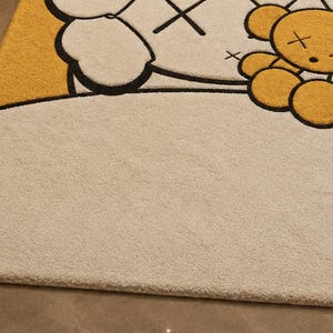 May include: A beige rug with a cartoon design. The rug features a large white character with black outlines and cross-shaped eyes. A yellow section and a smaller yellow bear-like character are also visible. The rug has a soft, textured surface.