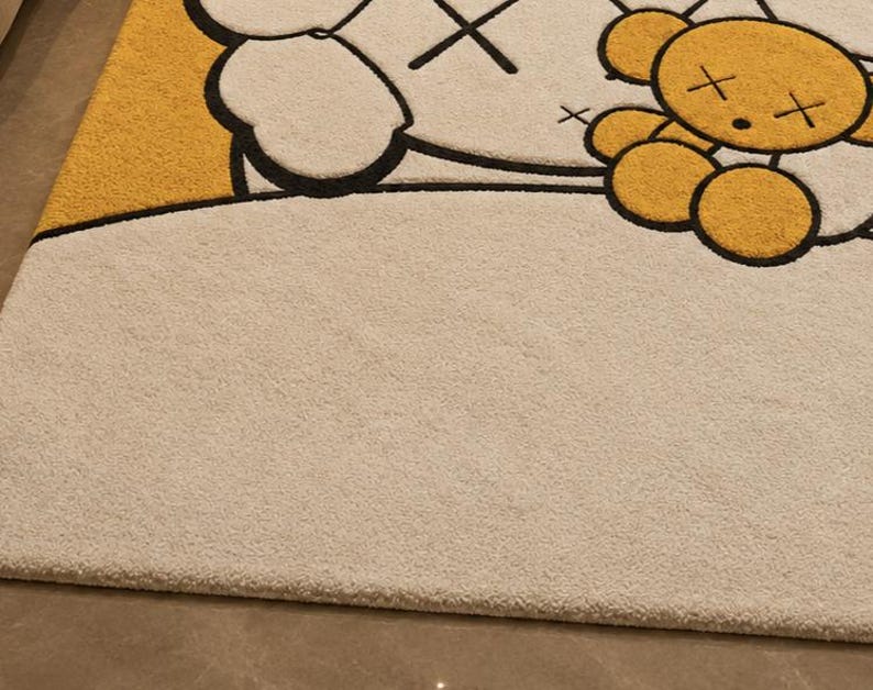 May include: A beige rug with a cartoon design. The rug features a large white character with black outlines and cross-shaped eyes. A yellow section and a smaller yellow bear-like character are also visible. The rug has a soft, textured surface.