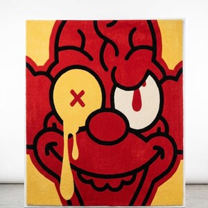 May include: A rectangular tufted rug featuring a stylized red clown face with a yellow background. The clown has a yellow eye with a red X and a dripping yellow liquid effect, a white eye with a red pupil, and a wide red smile outlined in black.