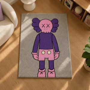 May include: A rectangular gray area rug featuring a centered cartoon character with a pink skull-shaped head, X-shaped eyes, and large ears. The character wears a purple long-sleeved shirt, pink shorts with two white buttons, and pink shoes.