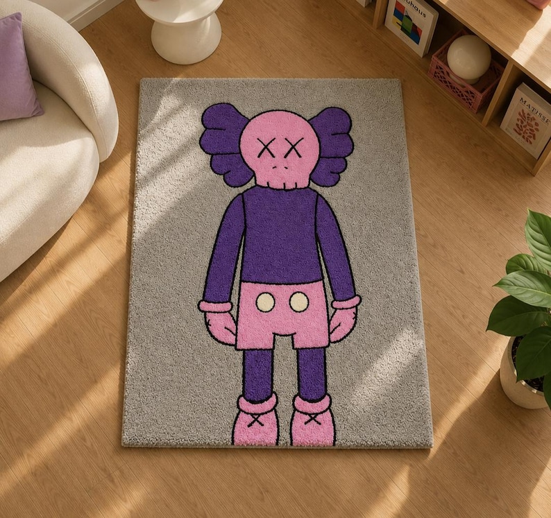 May include: A rectangular gray area rug featuring a centered cartoon character with a pink skull-shaped head, X-shaped eyes, and large ears. The character wears a purple long-sleeved shirt, pink shorts with two white buttons, and pink shoes.