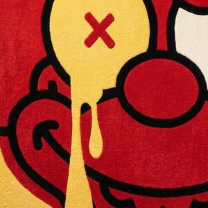 May include: A tufted rug featuring a stylized cartoon face in vibrant red and yellow. The design includes a prominent yellow eye with a red X, a dripping yellow effect, and bold black outlines that define the facial features and curved mouth.