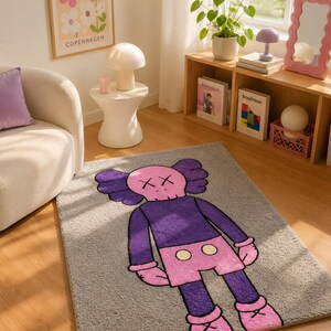 May include: A rectangular area rug featuring a gray background with a large, central graphic of a cartoon character. The character has a pink skull-shaped head with X-shaped eyes, a purple torso, pink shorts with two yellow circles, and pink shoes.