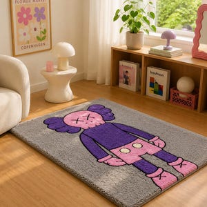 May include: A rectangular area rug featuring a gray background with a large, central graphic of a cartoon character. The character has a pink skull-shaped head with X-shaped eyes, purple ears, a purple torso, pink shorts with white buttons, and pink shoes.