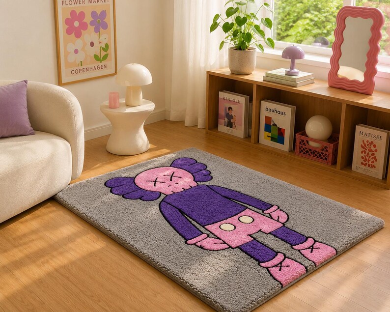 May include: A rectangular area rug featuring a gray background with a large, central graphic of a cartoon character. The character has a pink skull-shaped head with X-shaped eyes, purple ears, a purple torso, pink shorts with white buttons, and pink shoes.