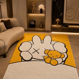 May include: A rectangular yellow and white rug featuring a cartoon character design. The rug has a large white character with black cross-shaped eyes and a small yellow teddy bear. The rug is placed on a light-colored floor.