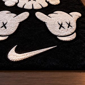 Pode incluir: Tapete de pel&uacute;cia preto e branco com um design de m&atilde;o de desenho animado, detalhes de olhos "XX" e um logotipo da Nike. O tapete tem uma superf&iacute;cie macia e texturizada, adequado para decora&ccedil;&atilde;o de casa.