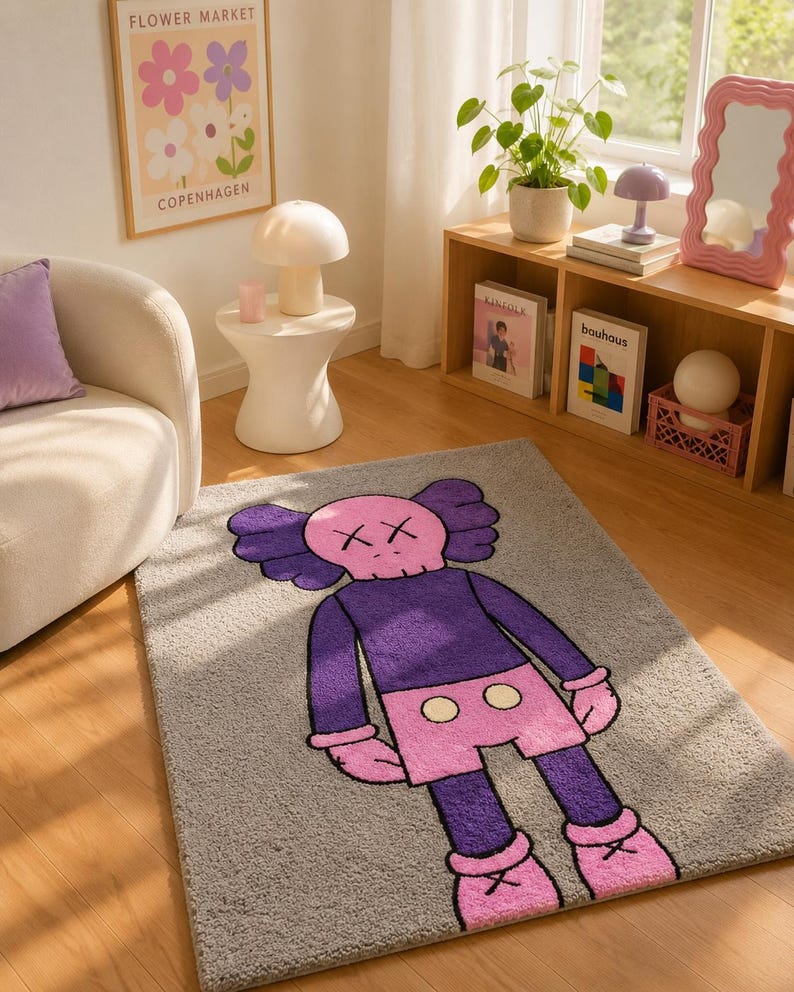 May include: A rectangular area rug featuring a gray background with a large, central graphic of a cartoon character. The character has a pink skull-shaped head with X-shaped eyes, a purple torso, pink shorts with two yellow circles, and pink shoes.
