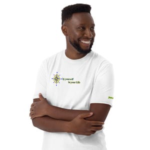Può includere: T-shirt bianca con un disegno a bussola e la scritta "by yourself in your Life" in verde e blu. La manica ha la scritta "Just a". La persona sorride.