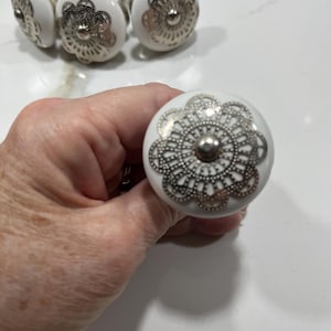 May include: A set of round white ceramic cabinet knobs featuring a decorative silver-toned metal filigree overlay in a floral pattern. Each knob has a smooth, glossy finish and a central silver-toned metal stud, providing a classic look.