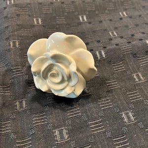 May include: A cream-colored ceramic drawer knob shaped like a blooming rose. The floral-shaped hardware features layered, curved petals with a smooth, glossy finish, designed to be mounted onto a piece of furniture or a cabinet door for functional use.