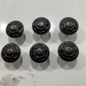 May include: A set of six round cabinet knobs in a dark, matte charcoal finish. Each knob features a smooth, raised circular center surrounded by a detailed, embossed scrollwork pattern that extends to the outer edge of the hardware piece.