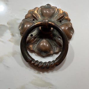May include: A circular copper-toned metal drawer pull featuring a decorative floral-shaped backplate with four mounting holes. The pull includes a hinged, twisted-texture ring handle that hangs downward from the center of the ornate metal base.