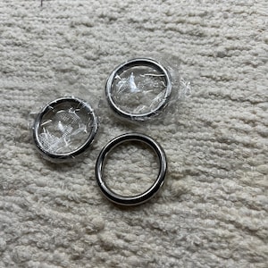May include: Three circular silver-toned metal rings with a smooth, polished finish. Two of the rings are individually encased in clear, crinkled plastic packaging, while the third ring is unwrapped and exposed, showing its solid metallic surface.