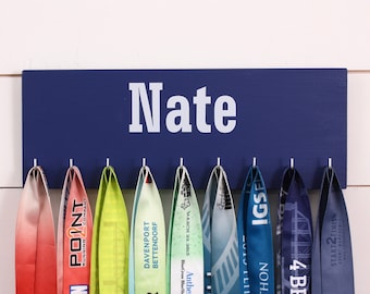 May include: A rectangular navy blue wooden medal display board with the name Nate printed in white capital letters. Eight silver metal hooks are attached to the bottom edge, each holding a colourful fabric race medal ribbon hanging vertically.