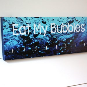 May include: A blue and black key holder with the text "Eat My Bubbles" printed in white. The key holder has eight silver hooks.