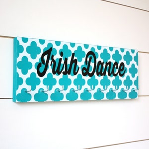 May include: A turquoise and white wooden sign with a repeating quatrefoil pattern and the text "Irish Dance" in black cursive lettering. The sign has eight silver hooks.