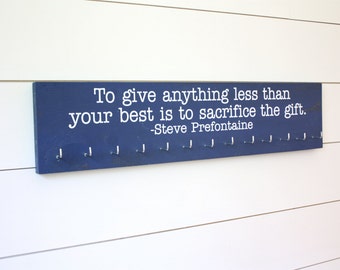 Könnte beinhalten: Ein rechteckiges, marineblaues Holzschild mit dem in Weiß aufgedruckten Zitat: To give anything less than your best is to sacrifice the gift. -Steve Prefontaine. Das Schild verfügt über eine Reihe von zwölf silbernen Metallhaken.