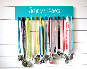 May include: A rectangular teal wooden medal display rack with the white text "Jennie's Races" printed on the front. The rack features twelve silver hooks, each holding a colourful fabric ribbon with a metal race medal attached at the bottom.
