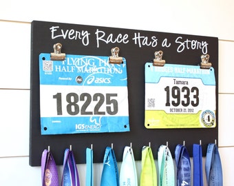 May include: Black chalkboard with the text "Every Race Has a Story" written in white chalk. Two race bibs are displayed with clips, one blue and one yellow. Below the bibs are eight colourful medals hanging on hooks.